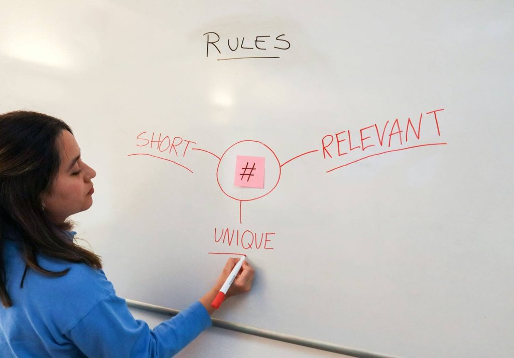 The image shows a person standing in front of a whiteboard, writing and engaging with the content on the board. The whiteboard contains a conceptual map or a set of rules that appear to revolve around the central theme denoted by a hashtag campaign symbol. The branches from the central theme point to words that describe characteristics these rules should possess: "SHORT," "RELEVANT," and "UNIQUE." These criteria suggest that the discussion is about creating effective hashtags or similar identifiers that are concise, pertinent, and distinctive. The person in the image seems to be in the process of writing or adding to the "UNIQUE" branch of the diagram, indicating active participation in formulating or understanding these rules.