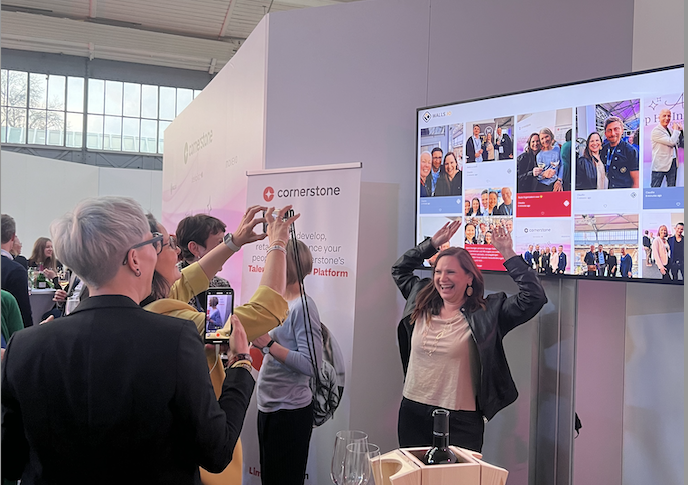 People are socializing and taking photos in front of a digital display wall at a professional event, where a woman is raising her hands at the HR influencers stage.