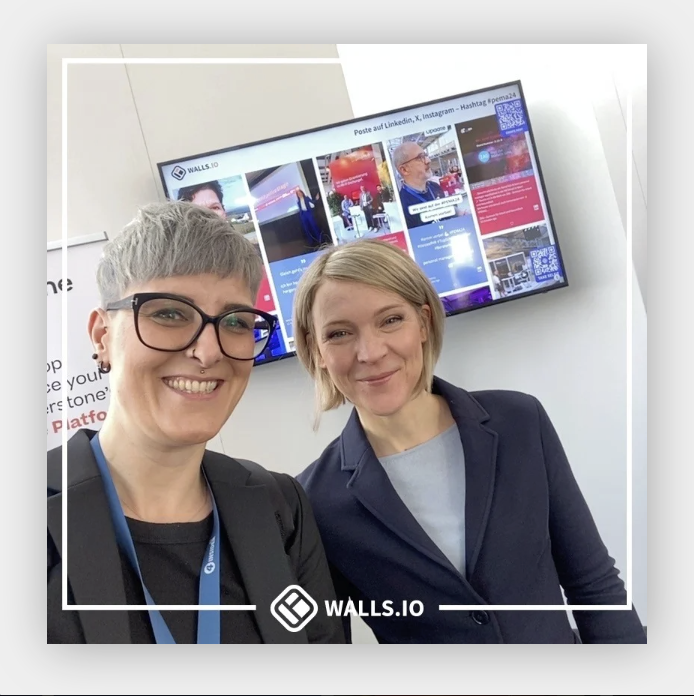 Two smiling individuals pose for a selfie in front of a digital display screen showing a social media wall from Walls.io.
