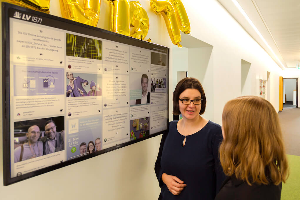 Two white people are chatting to each other in front of a large wall-mounted TV screen that shows the LV 1871 social wall. embed linkedin feed on a display