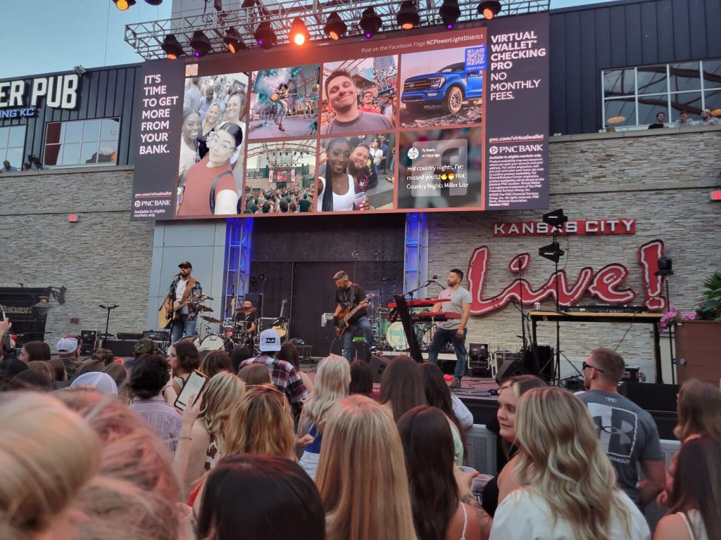 This image shows a lively outdoor concert scene at the "Kansas City Live!" venue, with a band performing on stage and a crowd of enthusiastic fans watching, while large screens display social media posts and event promotion material on the sides.