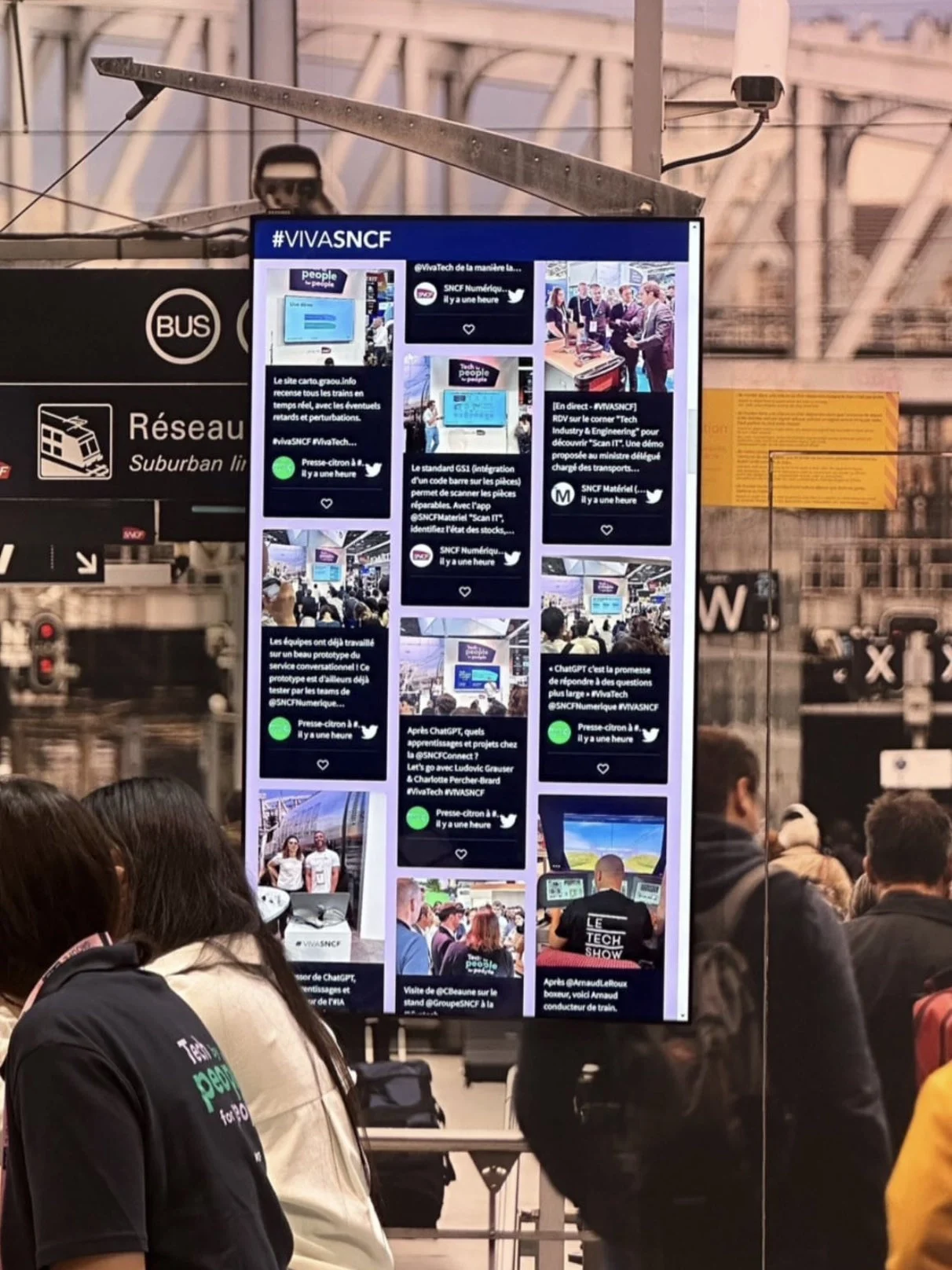 An experiantial events setup example. The image shows a digital screen at a trade show displaying a social media feed with the hashtag #VIVASNCF. The screen highlights various tweets related to SNCF (the French National Railway Company), focusing on their participation in the event, showcasing different activities, innovations, and interactions at the trade show. The background includes structural elements of the trade show venue, and there are signs related to transportation, such as "Suburban lines" and "BUS," suggesting SNCF's relevance in the context. Several people are gathered near the screen, engaged in conversation or exploring the trade show, reflecting a lively and interactive atmosphere.