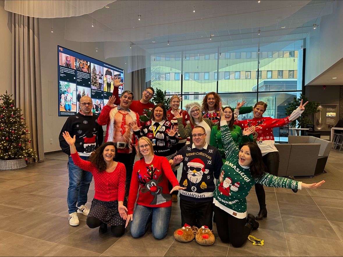 A group of colleagues dressed in festive Christmas sweaters pose cheerfully in an office lobby decorated with Christmas trees and holiday lights. They are smiling, raising their hands, and appear to be celebrating the holiday season.