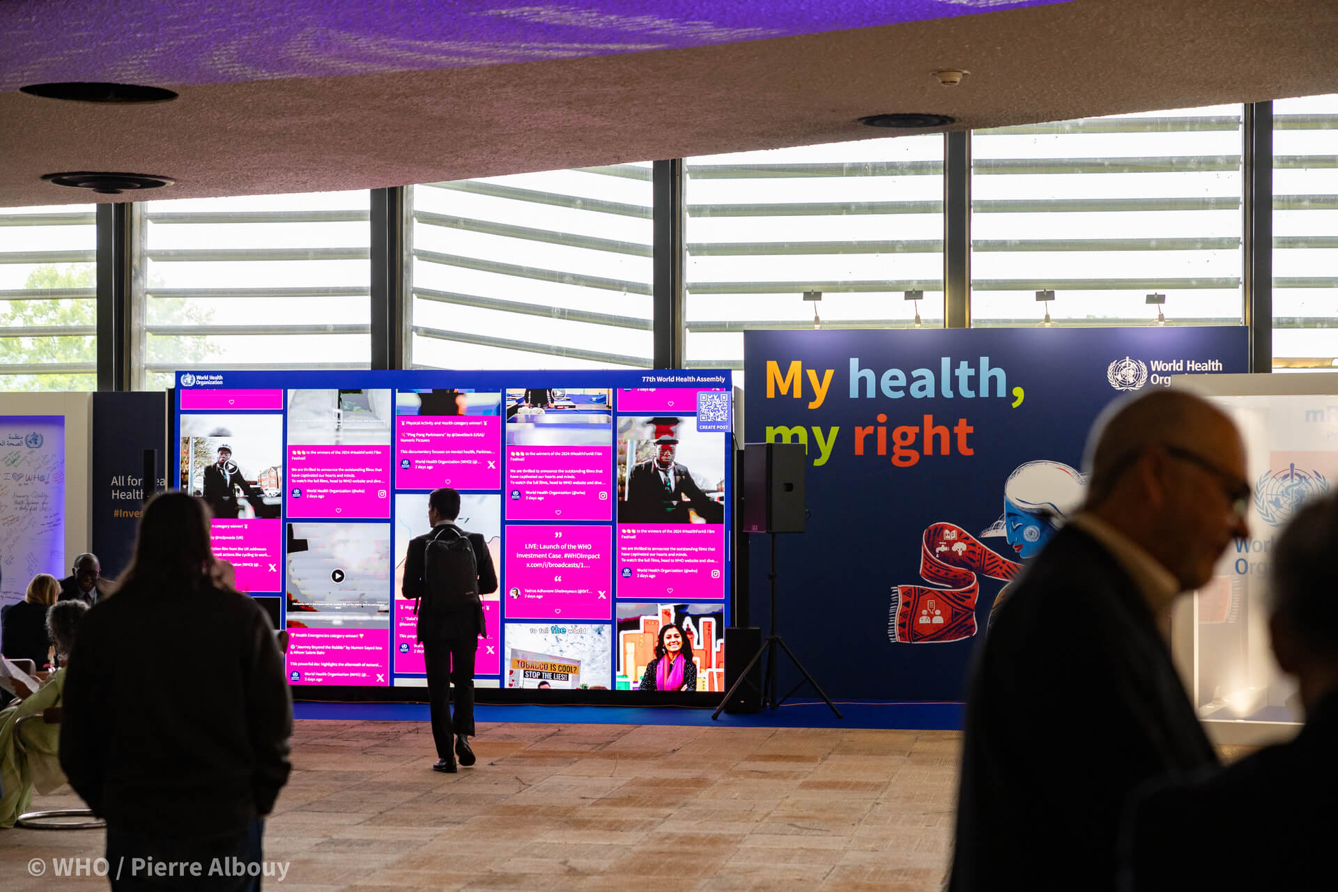 People are walking and interacting in a spacious lobby area, featuring large digital displays showing health-related content and banners, including one that states "My health, my right" at a World Health Organization event.