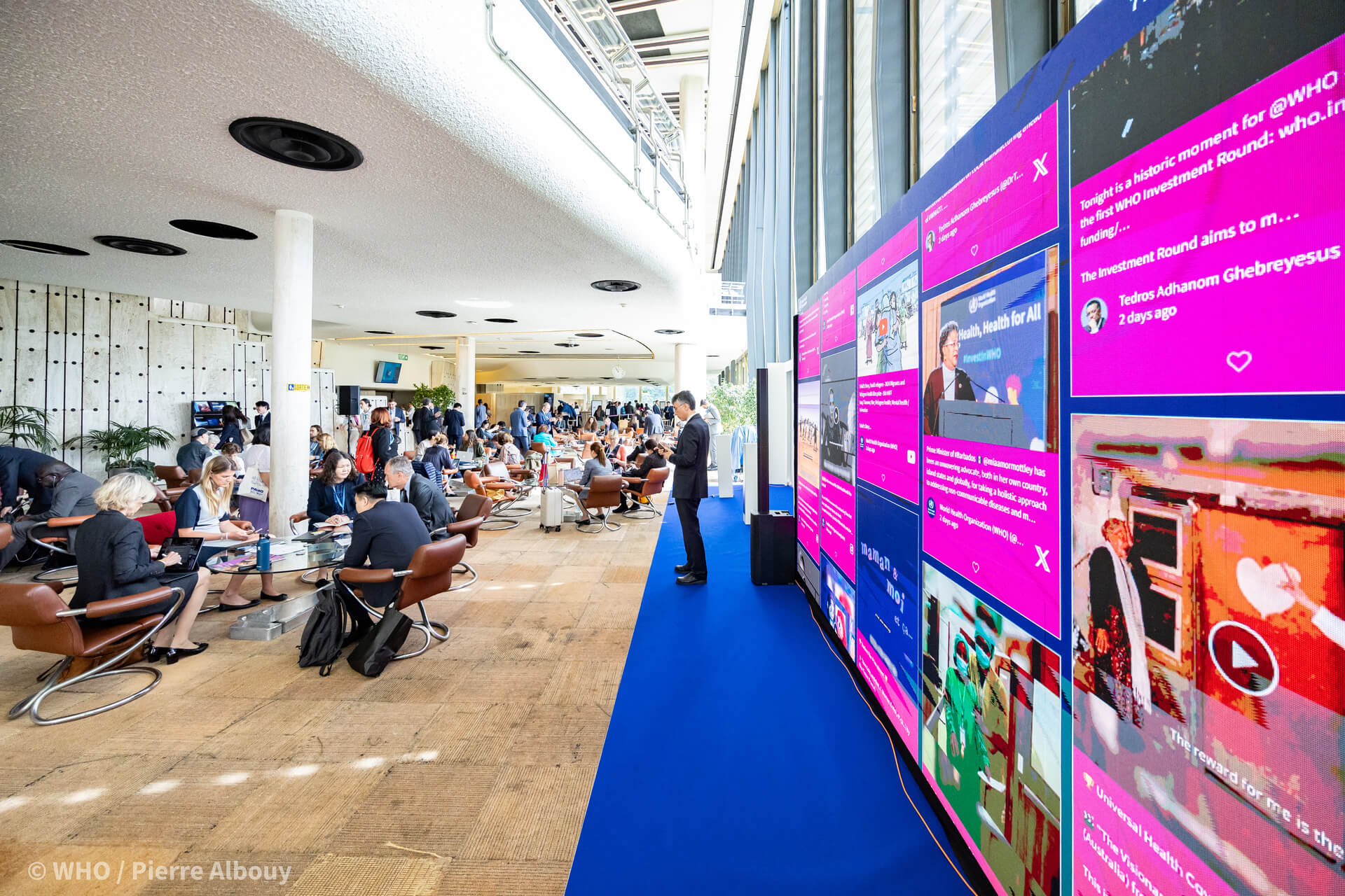 Attendees at a World Health Organization event are engaged in discussions and work on laptops in a lobby with large digital displays showcasing health-related content.