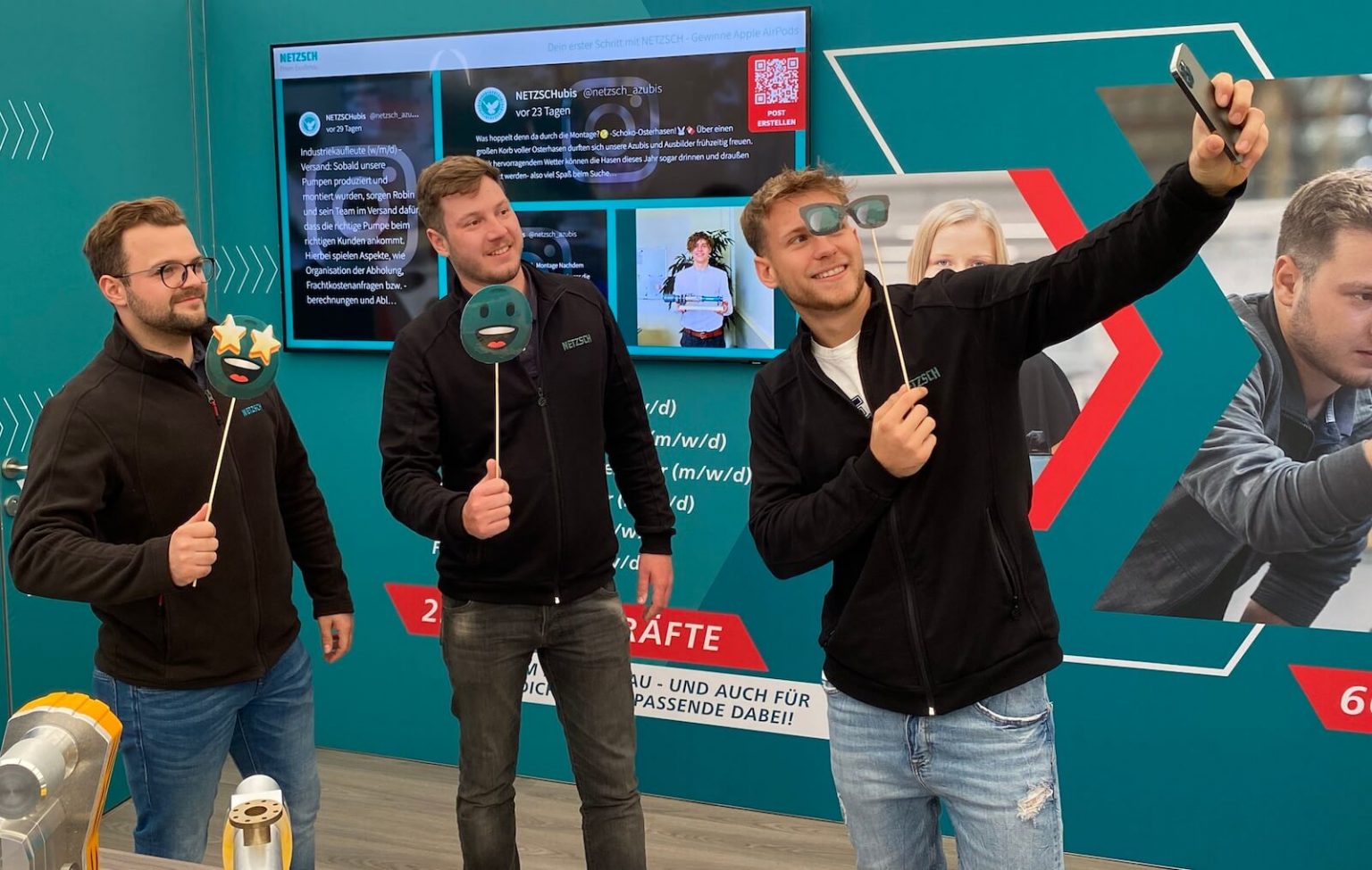 Three men using emoji props to take selfies in front of a corporate photo booth screen at an exhibition booth, enhancing engagement with visitors.