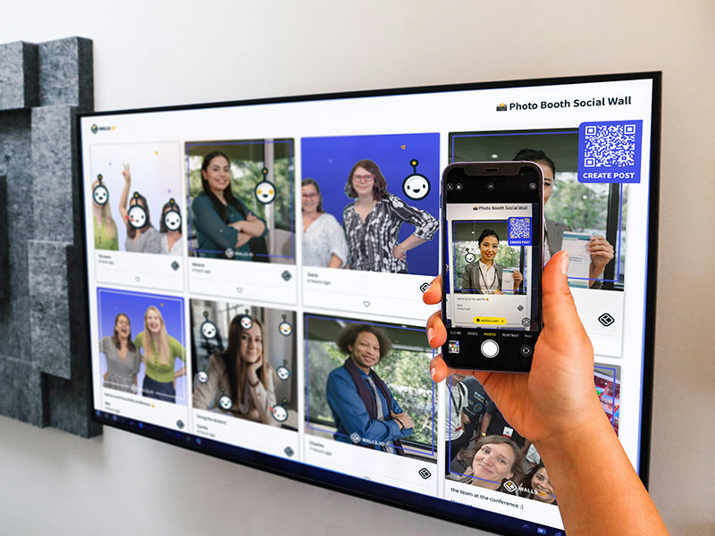 A hand holds a smartphone displaying a social wall post, while the same post is shown on a larger screen behind, creating a seamless digital experience for a photo booth at an event.